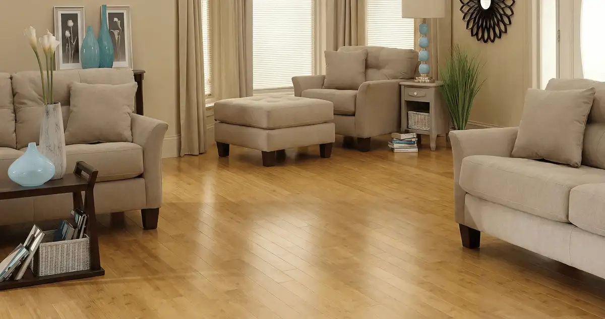 A living room with professionally installed bamboo flooring, beige sofas, a matching ottoman, a side table with a blue lamp, vases, framed pictures, and large windows with blinds and curtains in Norfolk, VA